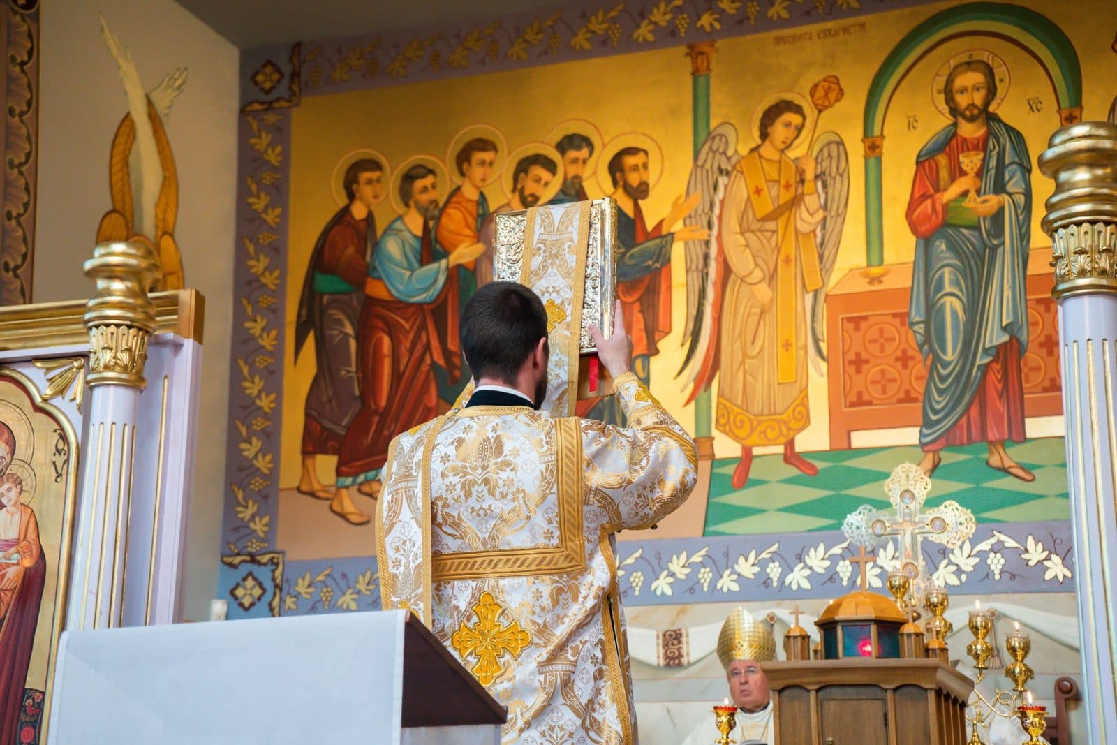 A vested minister raises the Gospel book before painted icons as another clergyman serves nearby.