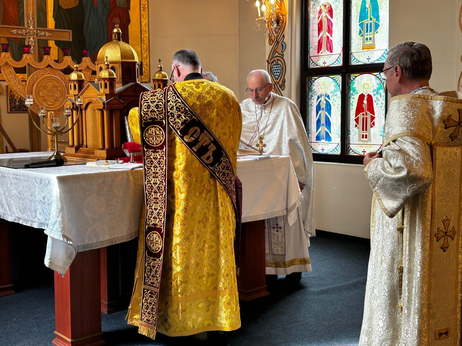 Clergy serve together around an altar beneath icons and stained-glass windows.
