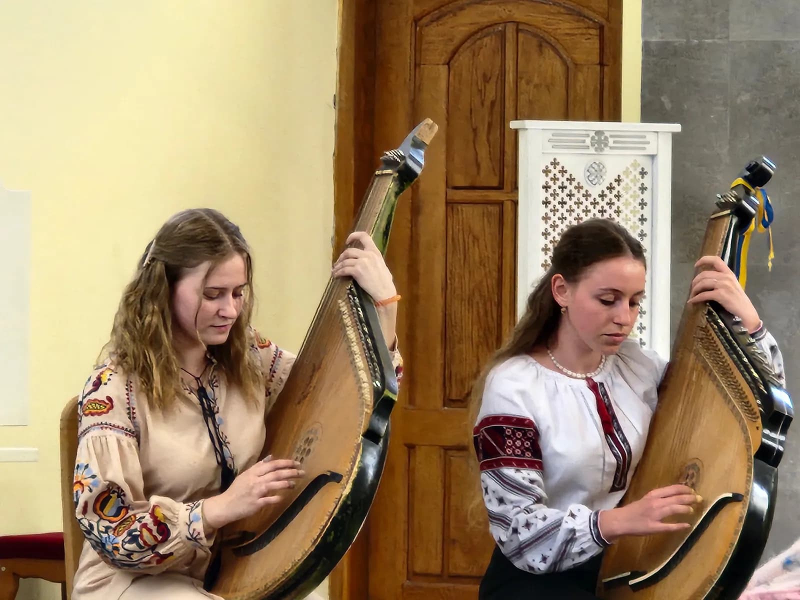 Two women in embroidered shirts play banduras in front of a wooden door.