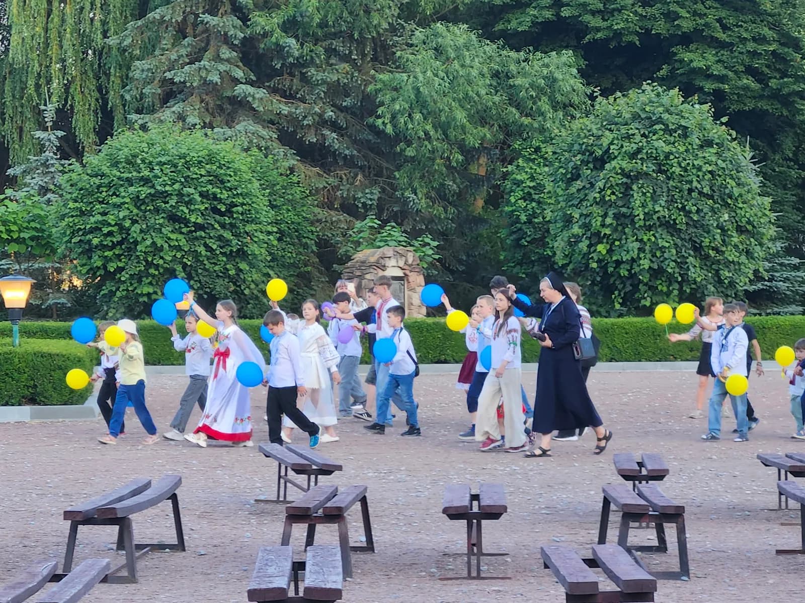 Children in traditional Ukrainian clothing hold blue and yellow balloons while walking with a nun.