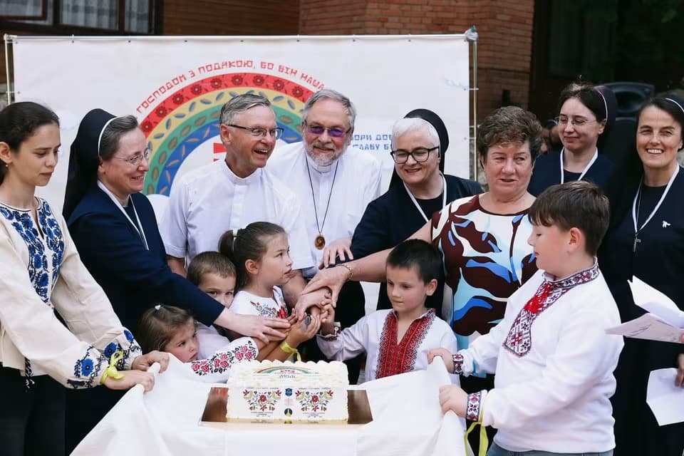 Clergy and children in embroidered shirts cut a decorated cake under a banner.