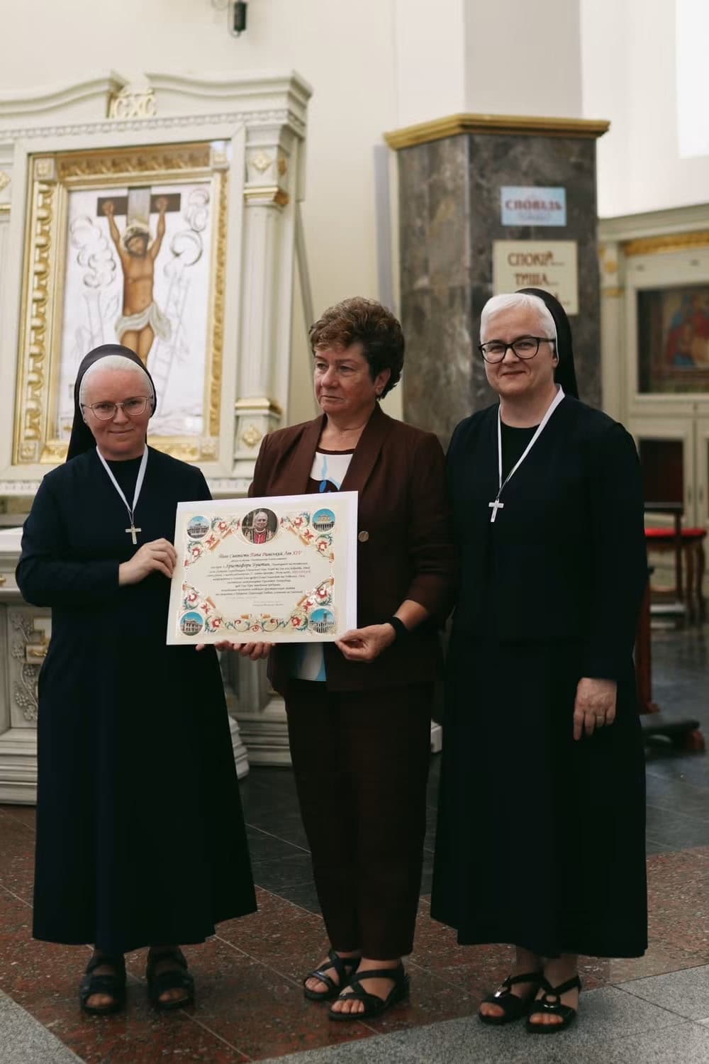 Three women in religious habits and a suit stand in a church holding a certificate.