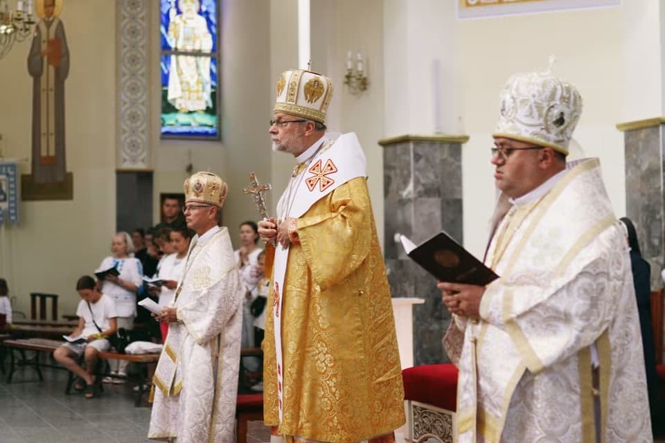Bishops in ornate vestments and mitres stand in church, one holding a processional cross.