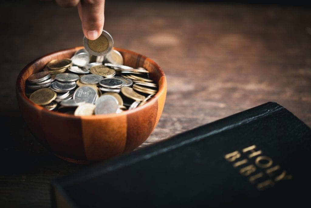 Hand drops coin into wooden bowl filled with coins next to Holy Bible.