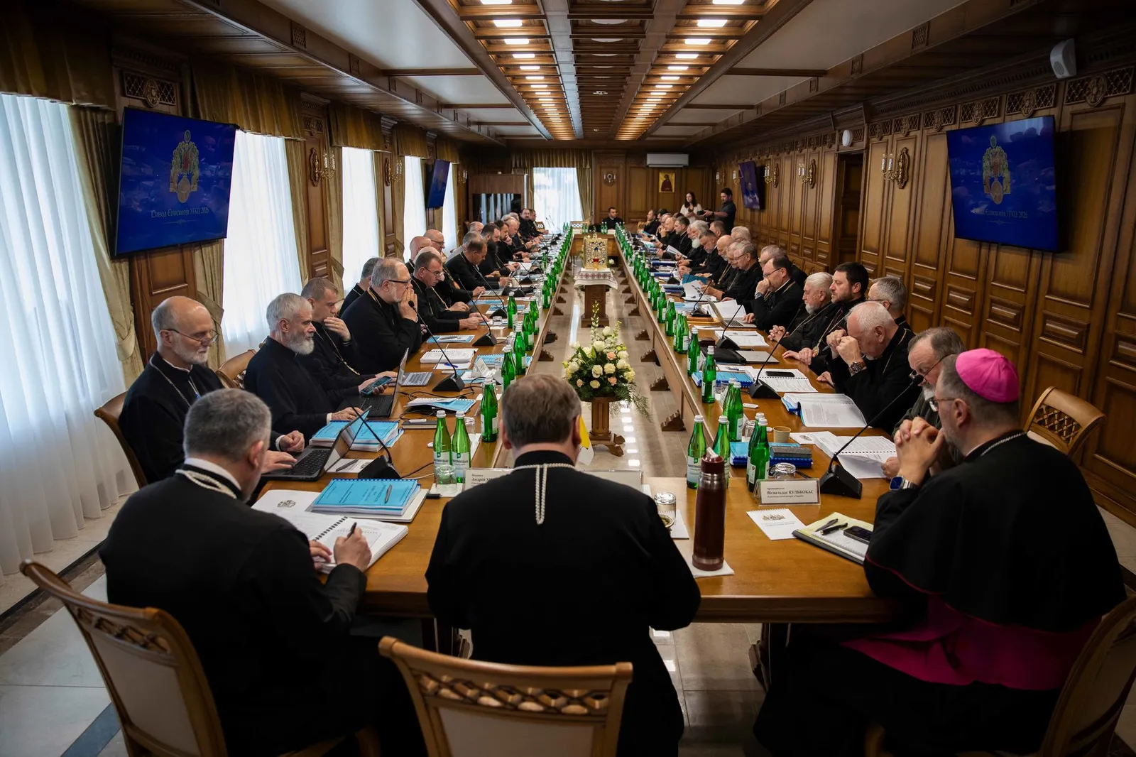Clergy gather around a long table for a meeting, with screens displaying church symbols.
