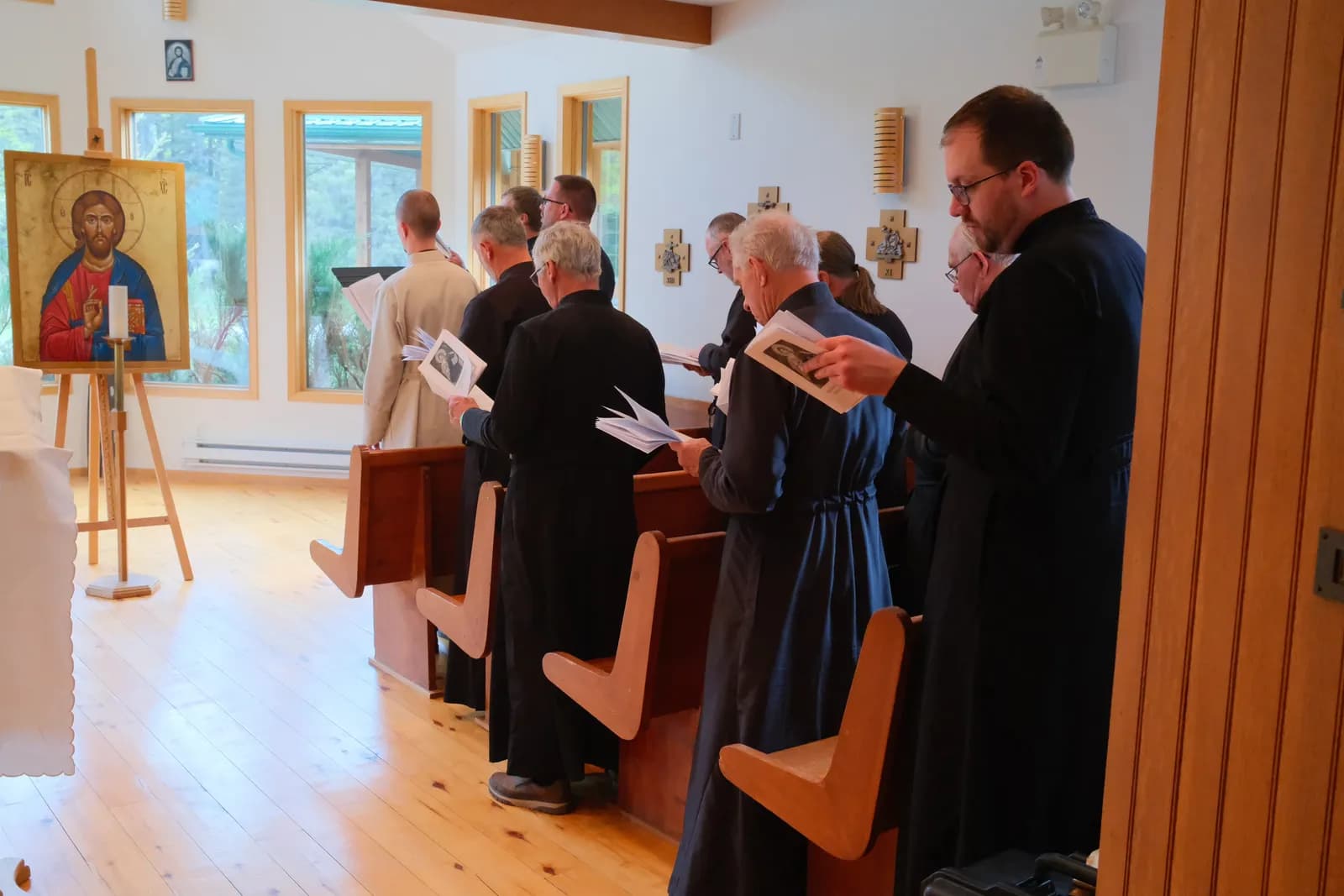 Clergy in cassocks stand in pews, reading from booklets, with an icon of Christ visible.