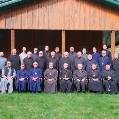 Clergy in cassocks and vestments pose for a group photo outside a wooden building.