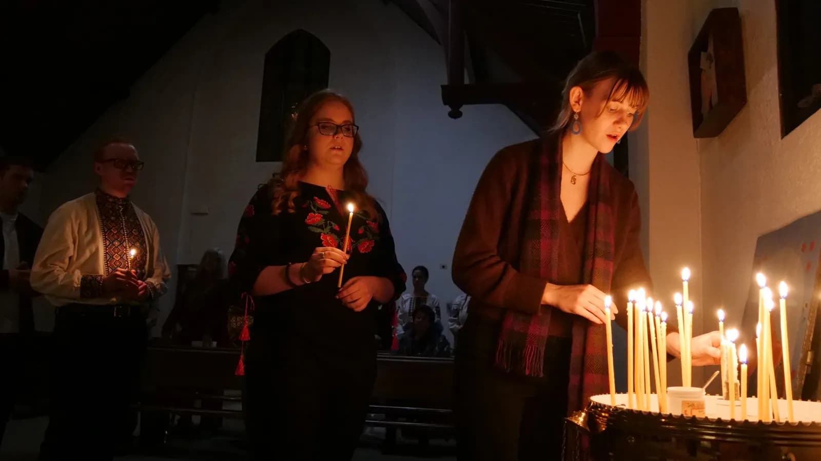 Young people light candles in a dimly lit church, some wearing embroidered shirts.