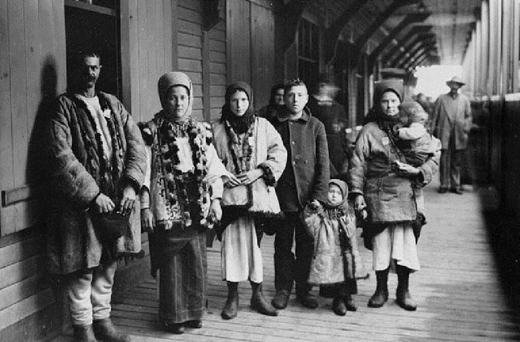 A group of immigrants in traditional clothing stand on a wooden platform.