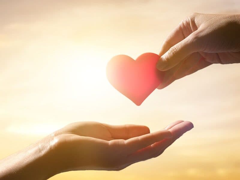 Two hands exchange a red heart against a bright, warm sky.