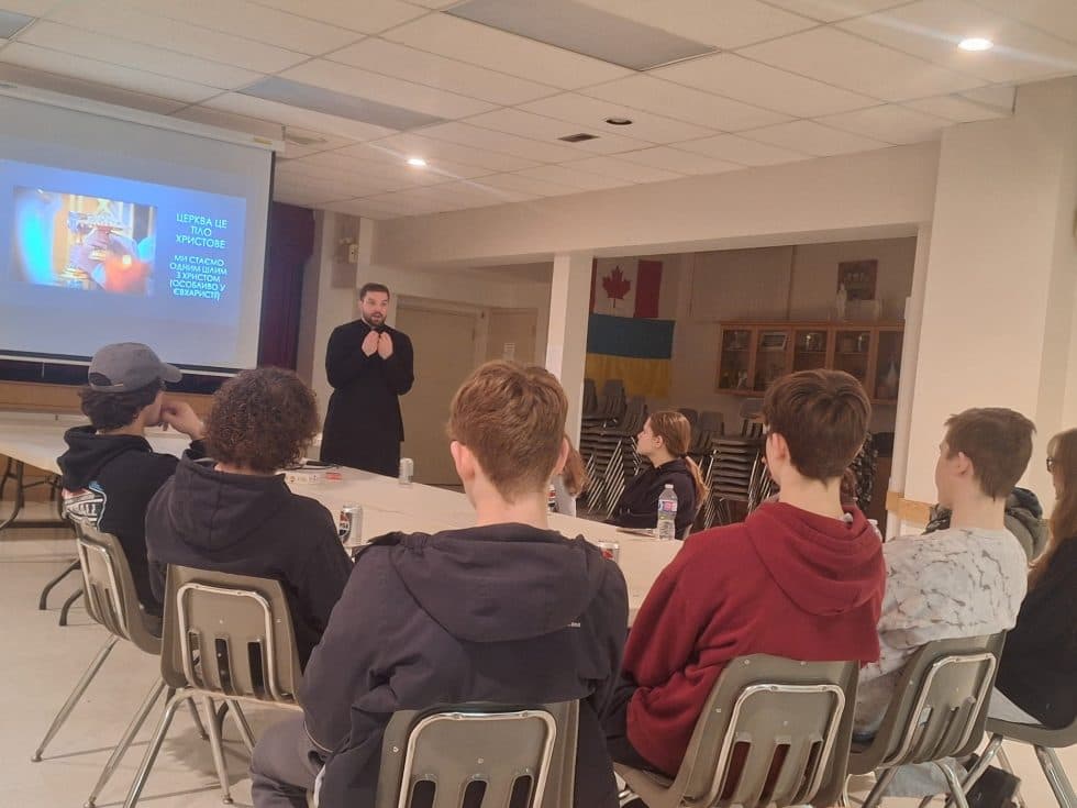 seminarian in cassock addresses youth group during presentation with Ukrainian and Canadian flags
