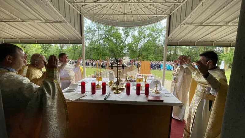 Clergy in vestments raise hands in prayer during an outdoor Divine Liturgy with a congregation gathered.