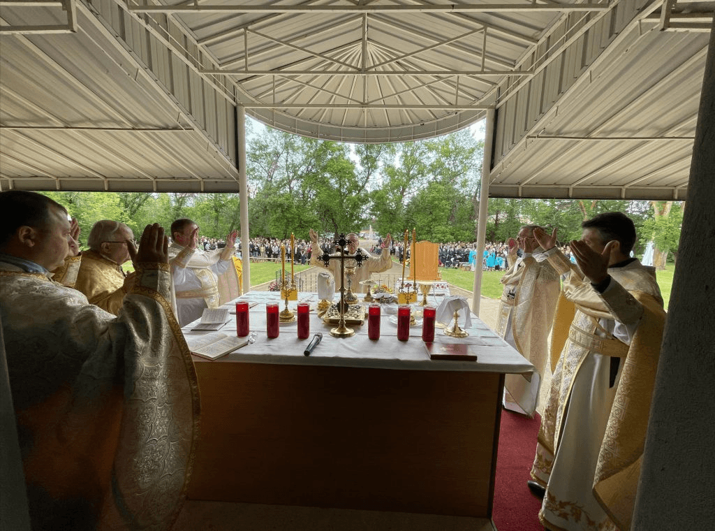 Clergy in vestments raise hands in prayer during an outdoor Divine Liturgy with a congregation gathered.