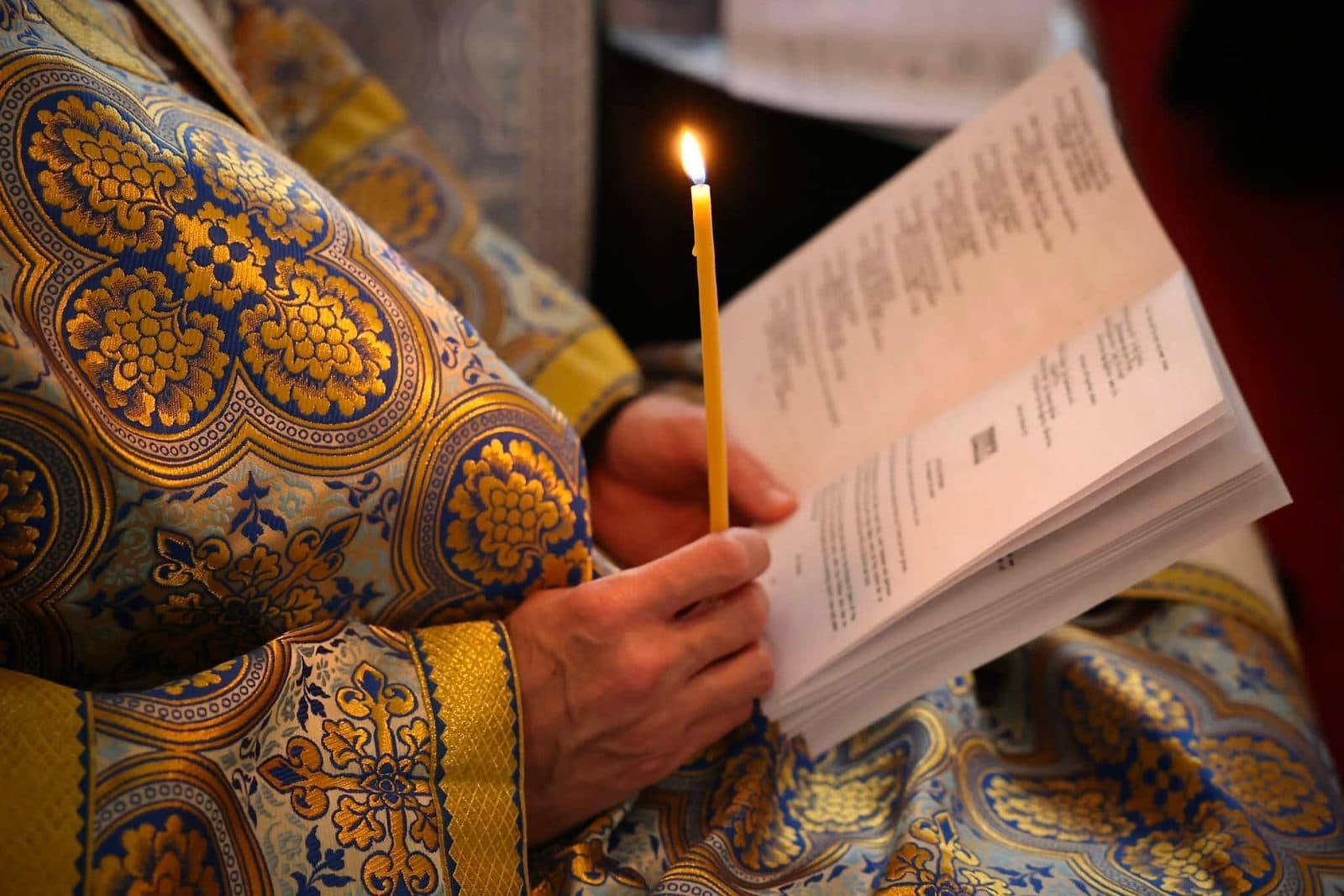 A priest reads from a liturgical book beside a candle in ornate blue vestments.