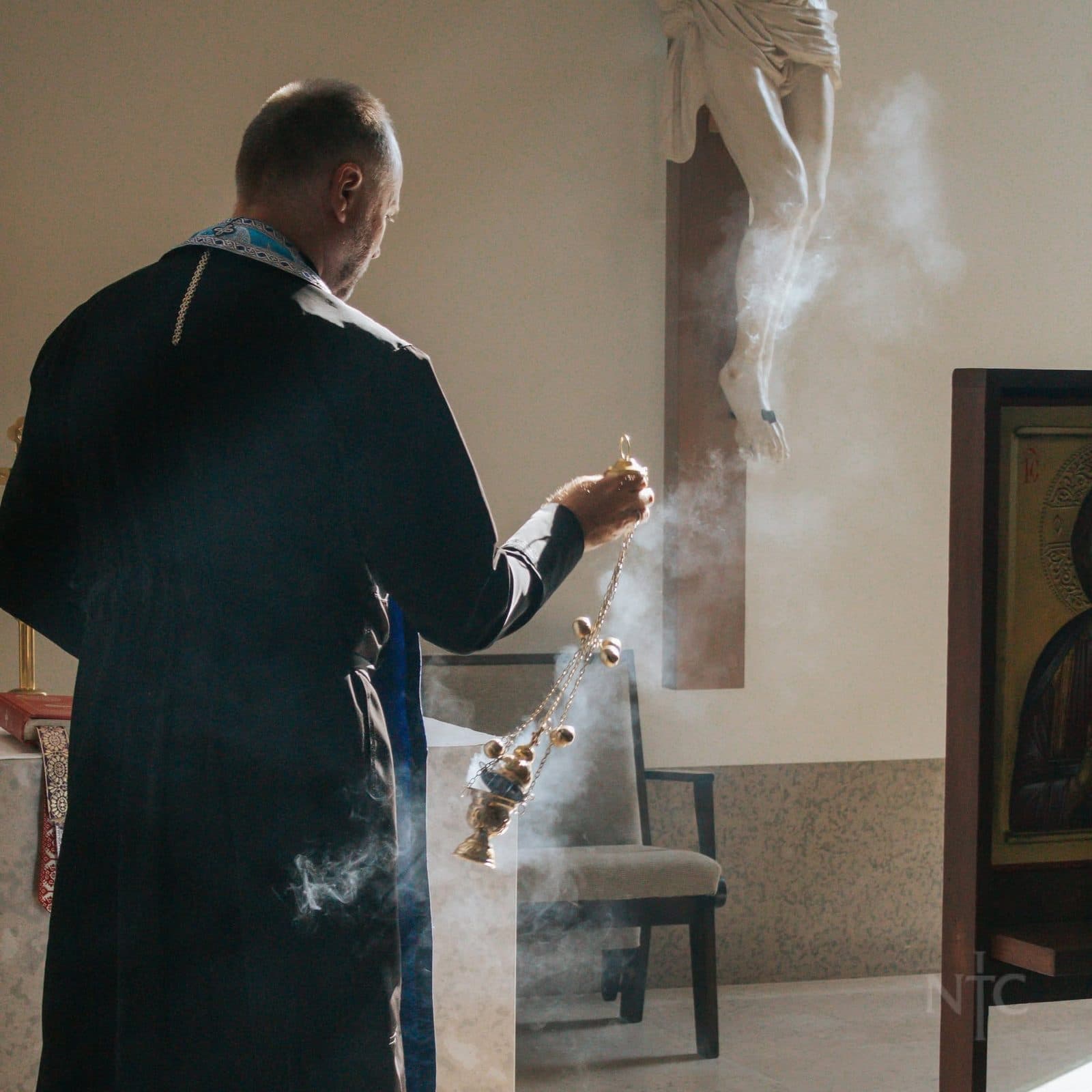 A priest in black vestments swings a censer beside a crucifix inside a church.