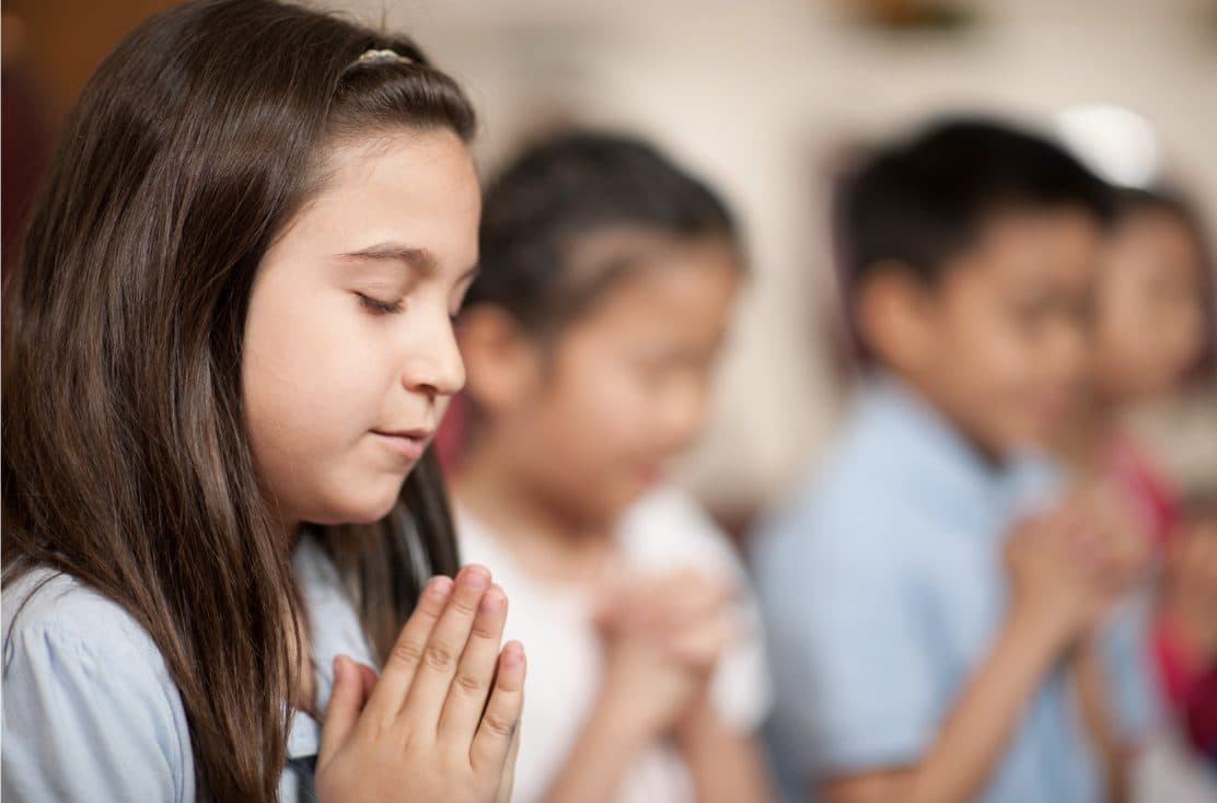 Children with eyes closed and hands clasped in prayer during a religious service.