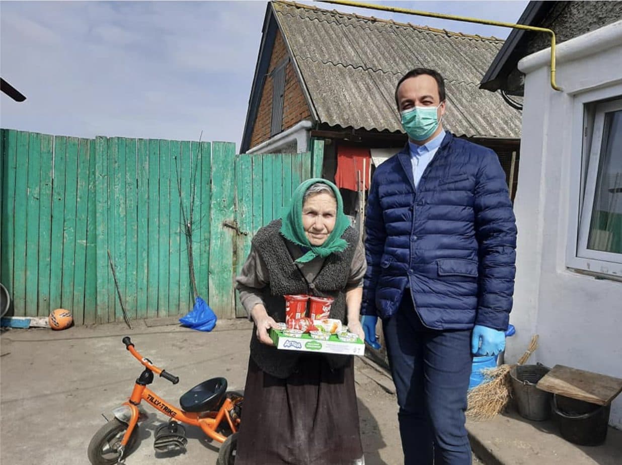 Elderly woman receives food donation from a masked man in a courtyard.