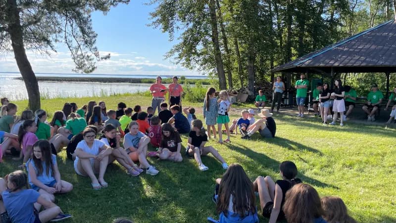 Children and adults gather outdoors by a lake for a camp activity.
