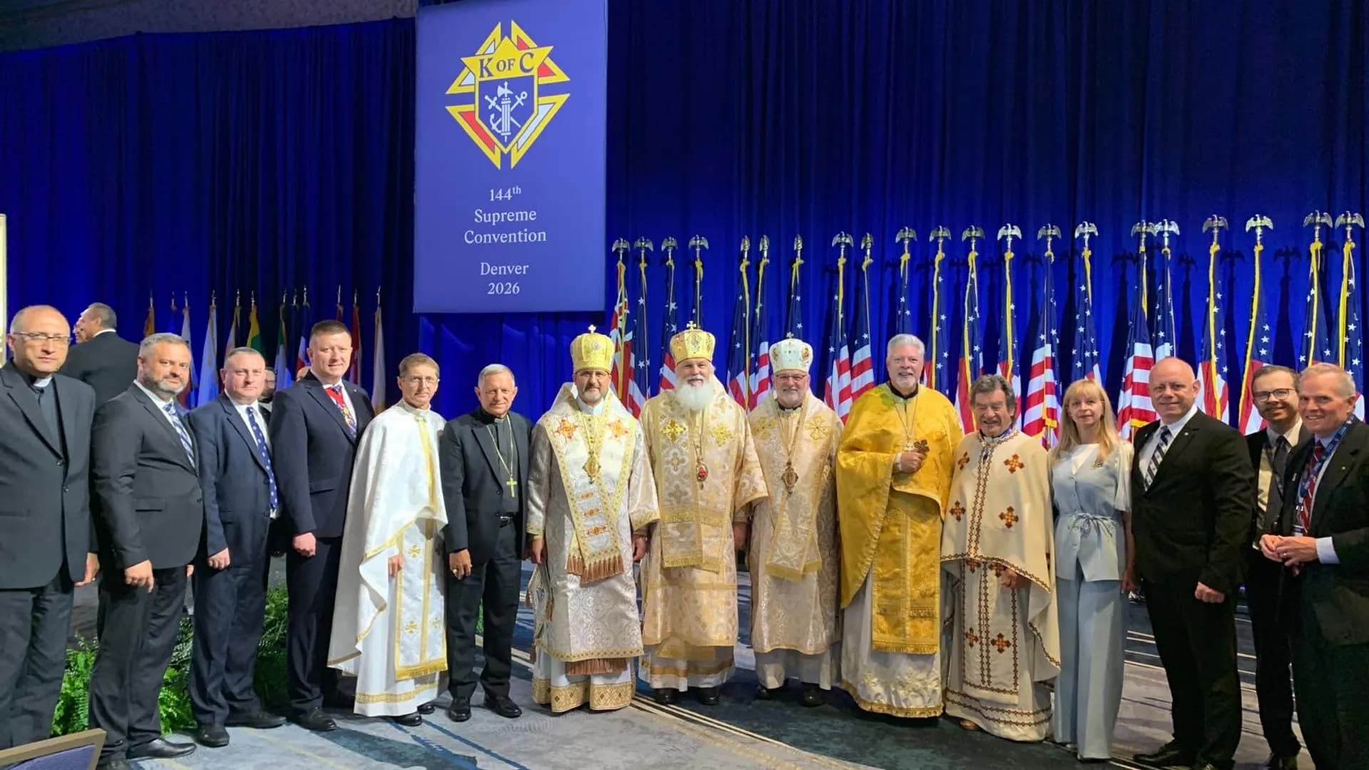 Clergy in liturgical vestments and laymen stand before a K of C banner