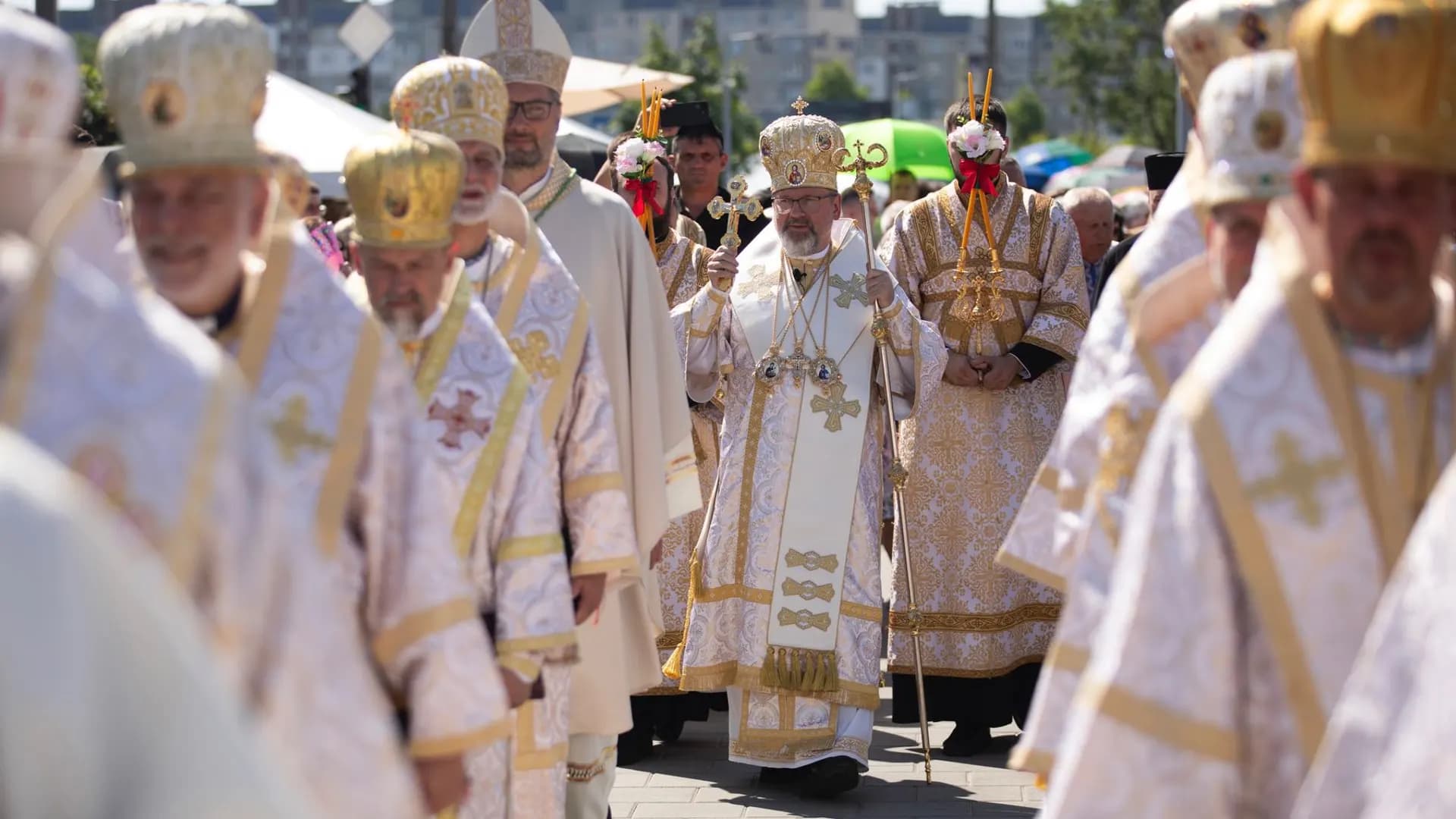 Clergy in ornate vestments and mitres process outdoors, led by a bishop holding a cross.